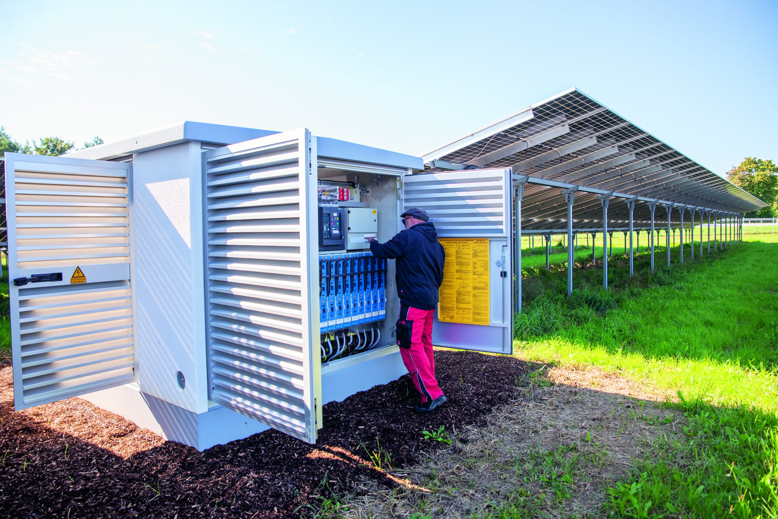 Techniker in schwarzer Jacke prüft Wechselrichter bei Solarpaneelen unter blauem Himmel.