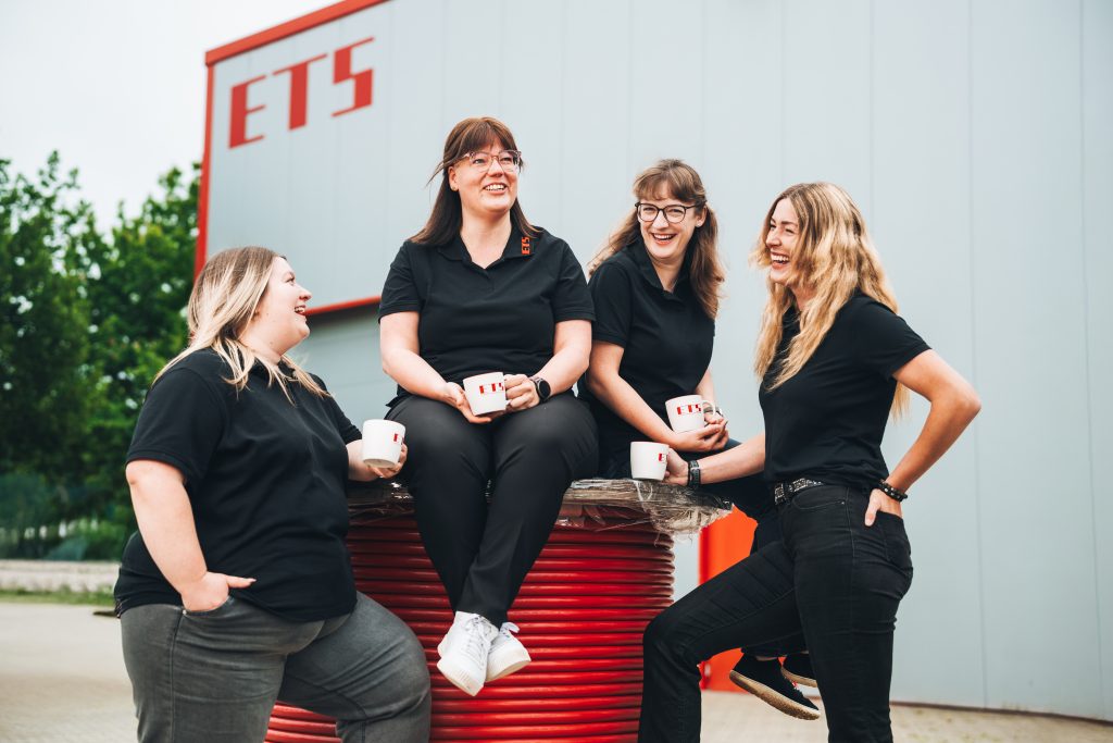Vier Frauen in schwarzen Polos plaudern lächelnd an roten Spulen im Industrieumfeld.