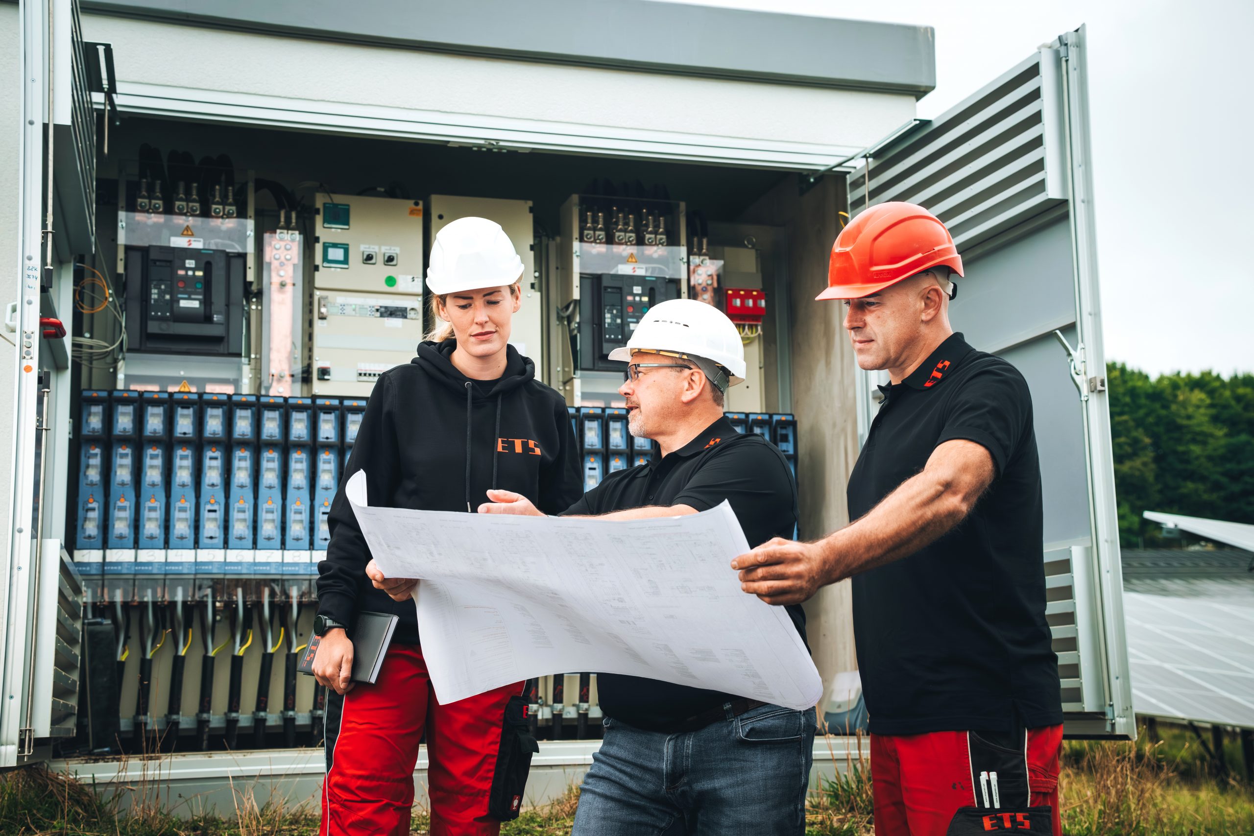 Drei Arbeiter mit Helmen besprechen Baupläne bei Solarpanels und elektrischer Ausrüstung