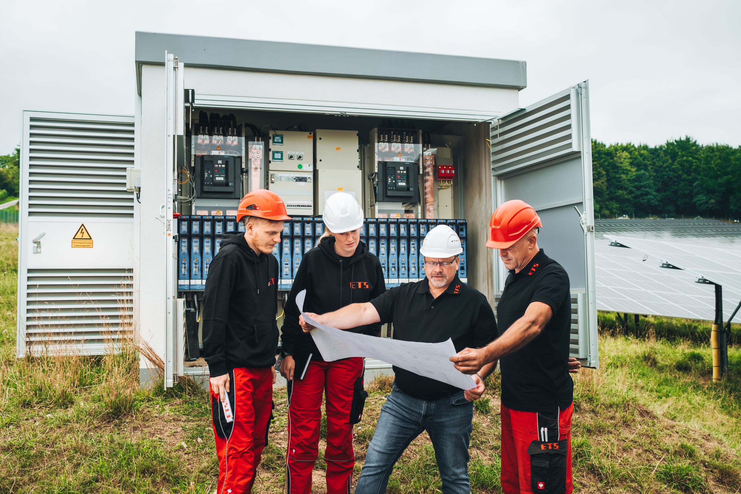 Vier Arbeiter mit Helmen prüfen Bauplan am Schaltschrank, Solarpaneele im Hintergrund.