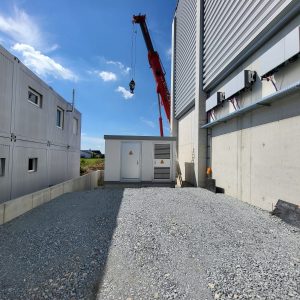 A crane lifts equipment near a construction site, with gravel ground and industrial buildings under a bright blue sky.