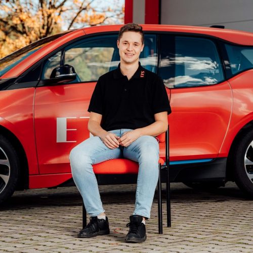 Jonas Reisinger sitzt vor rotem E-Auto in schwarzem Polo und Jeans, Hintergrund unscharf.