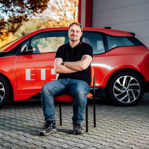 Maximilian Säurig sitzt vor rotem E-Auto in schwarzem Polo und Jeans, Hintergrund unscharf.