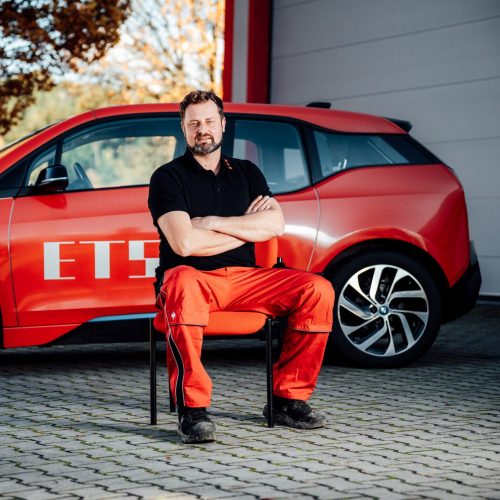 Juri Kostin sitzt vor rotem E-Auto in schwarzem Polo und roter Arbeitshose, Hintergrund unscharf.