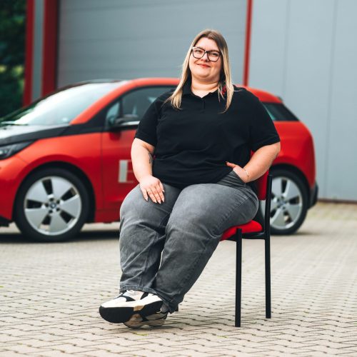 Katrin Menges​ sitzt vor rotem E-Auto in schwarzem Polo und Jeans, Hintergrund unscharf.