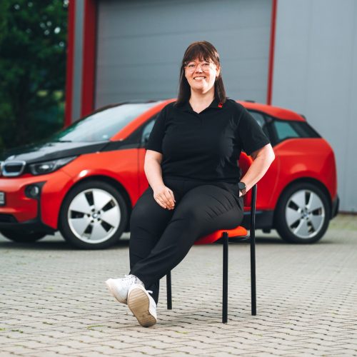 Kerstin Völkl sitzt vor rotem E-Auto in schwarzem Polo und Jeans, Hintergrund unscharf.