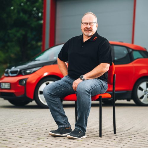 Josef Attenberger sitzt vor rotem E-Auto in schwarzem Polo und Jeans, Hintergrund unscharf.