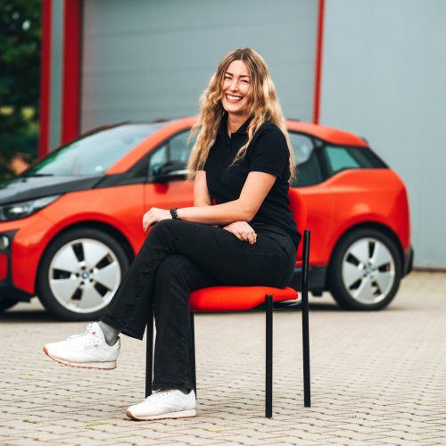 Theresa Vogl​ sitzt vor rotem E-Auto in schwarzem Polo und Jeans, Hintergrund unscharf.