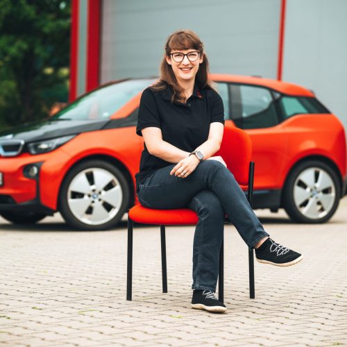 Lisa Gmeinwieser​ sitzt vor rotem E-Auto in schwarzem Polo und Jeans, Hintergrund unscharf.