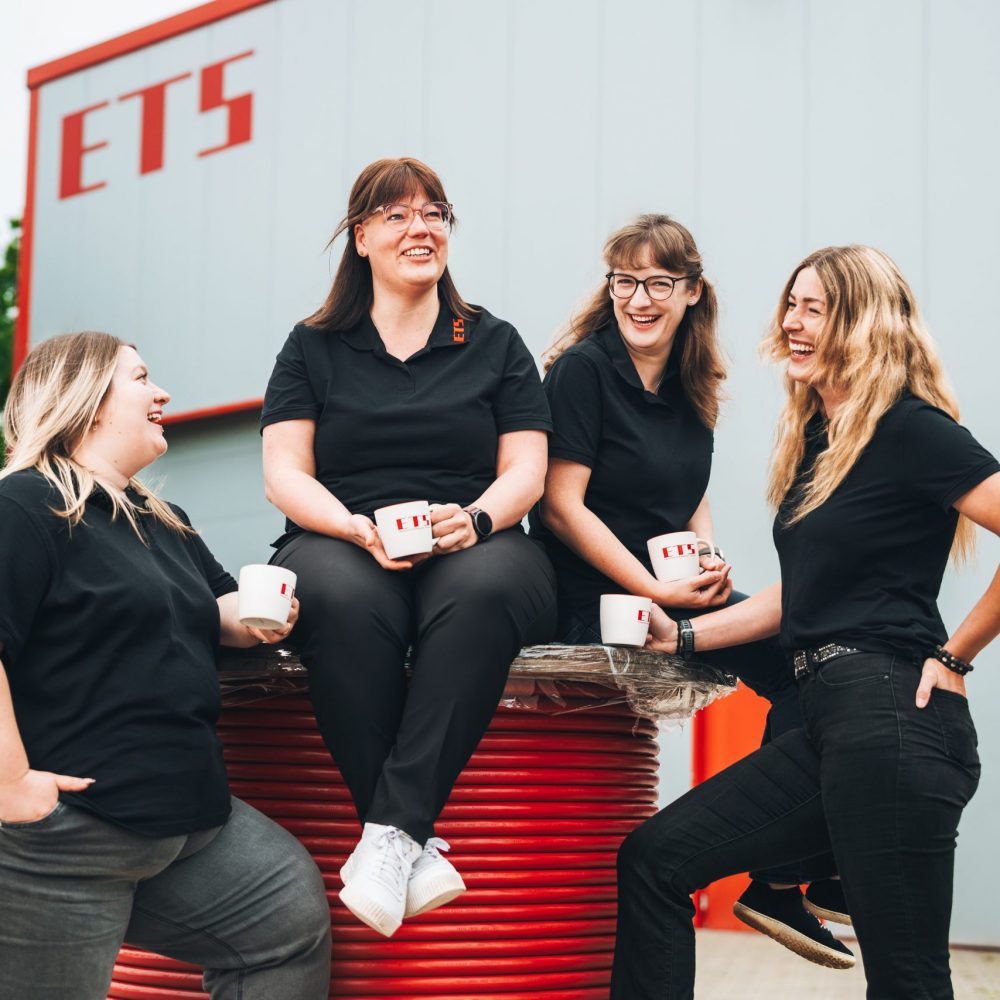 Vier Frauen in schwarzen Polos plaudern lächelnd an roten Spulen im Industrieumfeld.