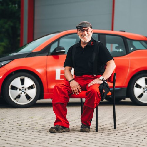 Alexander Aich​ sitzt vor rotem E-Auto in schwarzem Polo und Jeans, Hintergrund unscharf.