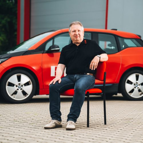 André Gölzer sitzt vor rotem E-Auto in schwarzem Polo und Jeans, Hintergrund unscharf.