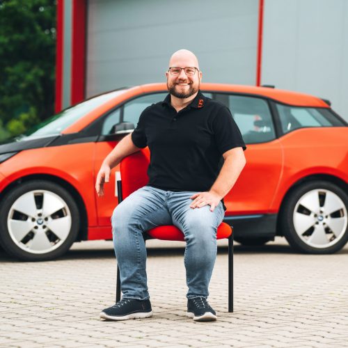 Marcus Grassl sitzt vor rotem E-Auto in schwarzem Polo und Jeans, Hintergrund unscharf.