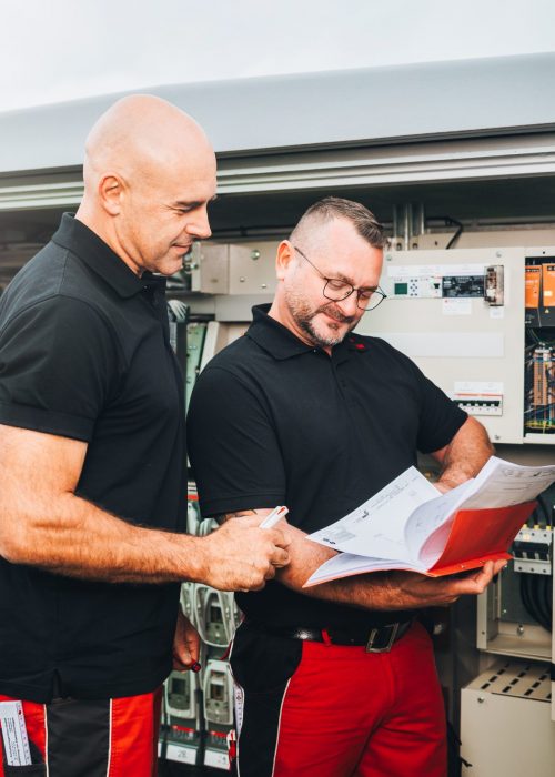 Zwei Techniker in schwarzen Shirts prüfen ein Handbuch bei elektrischen Schaltschränken