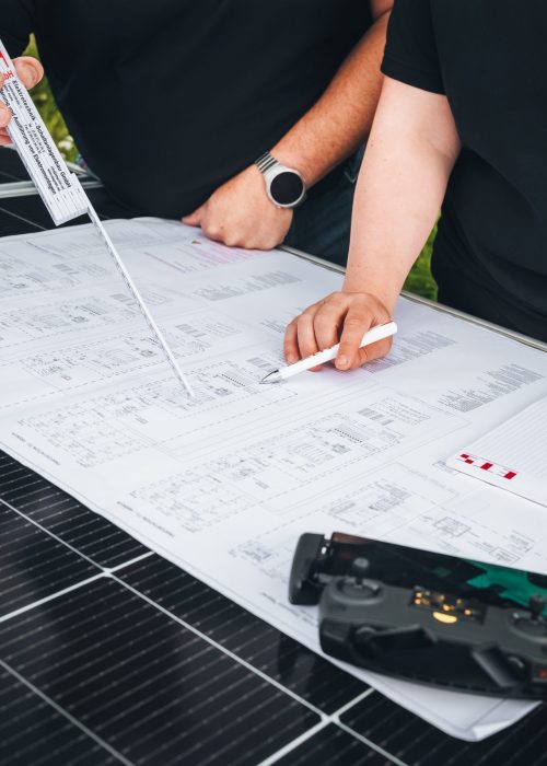 Zwei Personen in schwarzen Shirts prüfen technische Pläne auf Solarpanel mit Messwerkzeug